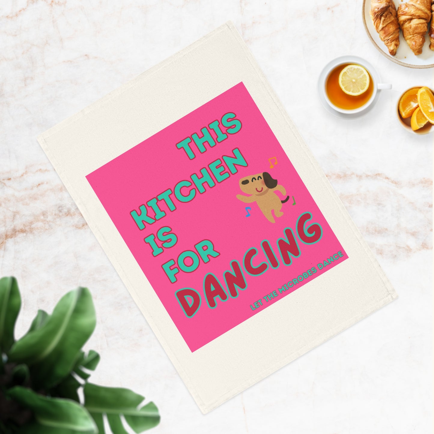 Nahaufnahme eines 'This Kitchen is for Dancing'-Küchentuchs auf einer Marmoroberfläche, dekoriert mit Pflanzen und Frühstückszubehör.