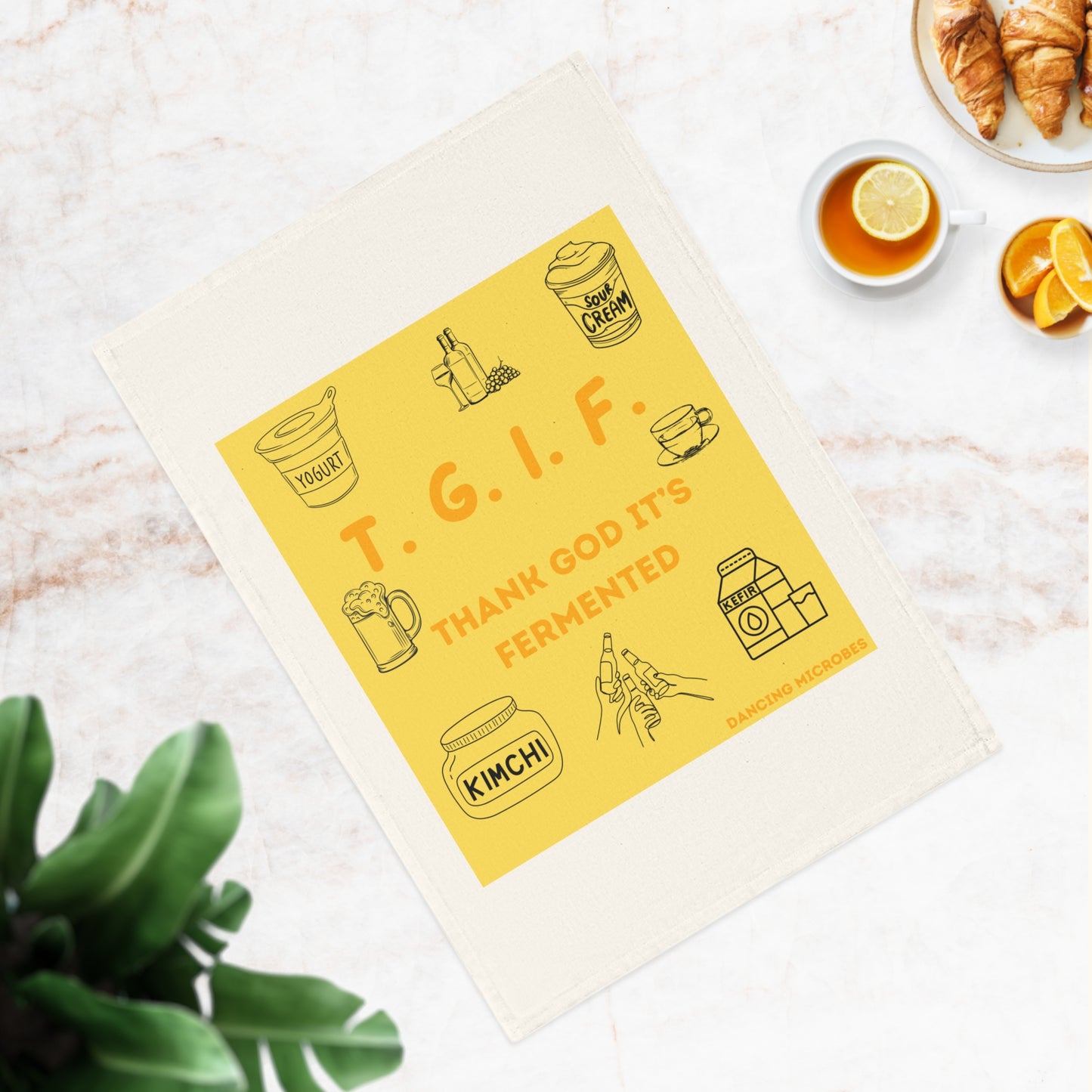 Nahaufnahme eines 'T.G.I.F.'-Küchentuchs auf einer Marmoroberfläche, dekoriert mit grünen Pflanzen und Essensaccessoires.