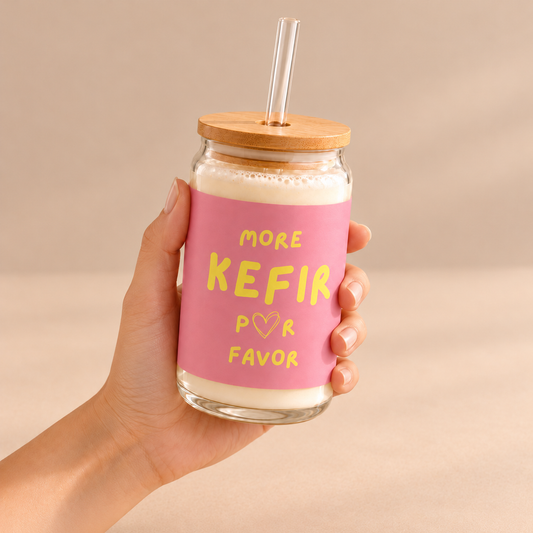 Eine Hand hält das mit Milchkefir gefüllte More Kefir Por Favor Trinkglas mit rosa Aufdruck, Bambusdeckel und Trinkhalm.