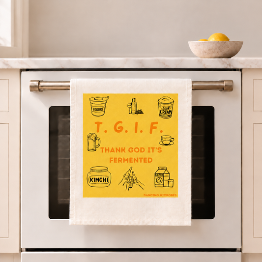 Weißes TGIF-Geschirrtuch von Dancing Microbes mit gelbem Motiv und der Aufschrift „Thank God It’s Fermented“, aufgehängt am Backofengriff.