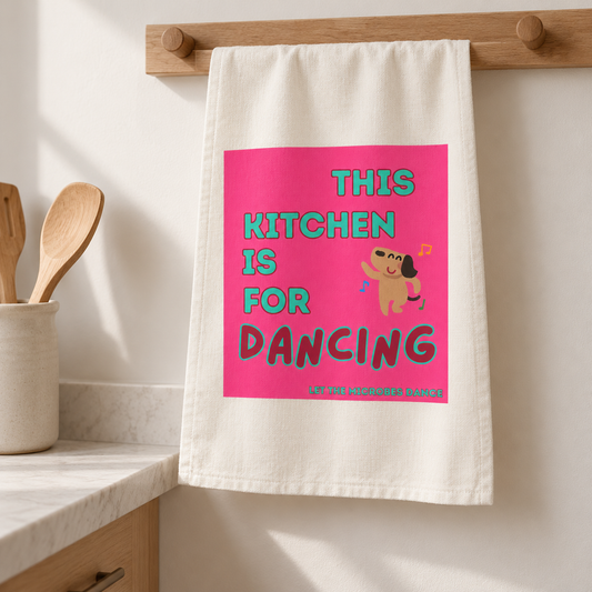 Cremefarbenes Geschirrtuch von Dancing Microbes mit pinkem „This Kitchen Is for Dancing“-Aufdruck, aufgehängt an einer Holzleiste in einer hellen Küche.