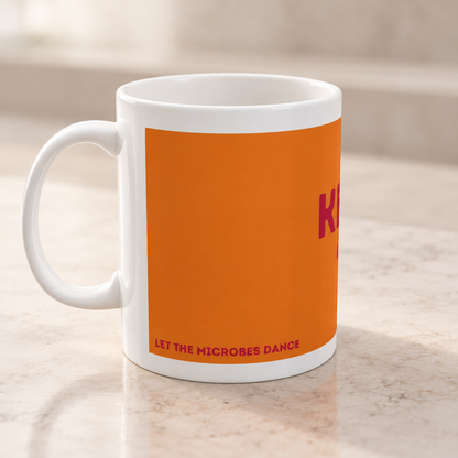 Orangefarbene Keramiktasse von Dancing Microbes mit weißem Henkel links und teilweise sichtbarem Schriftzug auf einer hellen Arbeitsfläche.