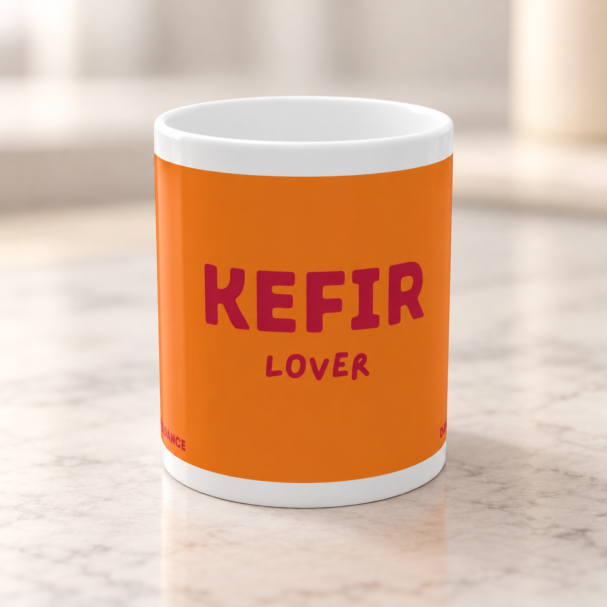 Orangefarbene Kefir Lover Tasse mit dunkelrosa Schriftzug auf einer hellen Arbeitsfläche vor einem weich verschwommenen Hintergrund.