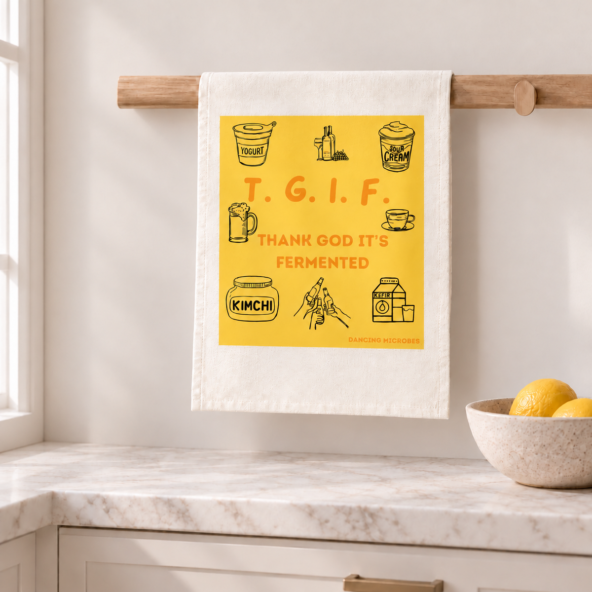 Weißes Geschirrtuch mit gelbem TGIF-Print und kleinen Fermentationsillustrationen, aufgehängt an einer Holzleiste in einer hellen Küche.