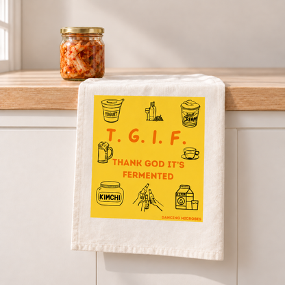 Weißes TGIF-Geschirrtuch mit gelbem „Thank God It’s Fermented“-Aufdruck, aufgehängt an einer Holzleiste in einer modernen Küche.